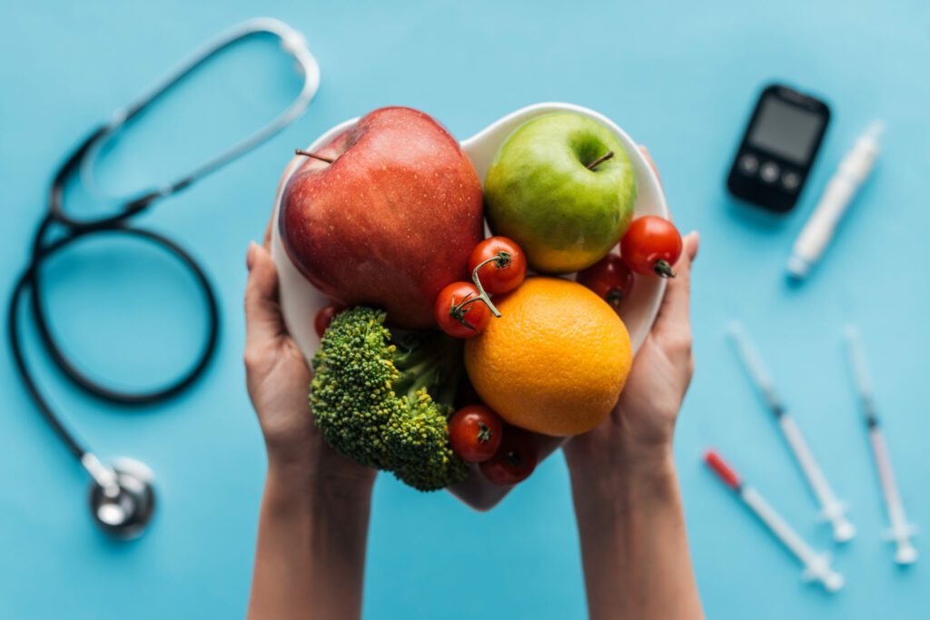 Two hands holding a heart-shaped bowl of fresh fruit and vegetables over a blurred background of medical equipment