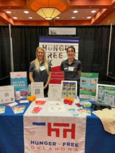Two HFO staff members stand with the HFO booth at the Oklahoma Libraries Association conference in March 2023