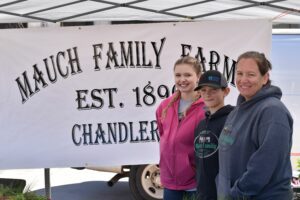 Three people standing in front of a sign that says Mauch Family Farm, Est. 189 (date obscured), Chandler